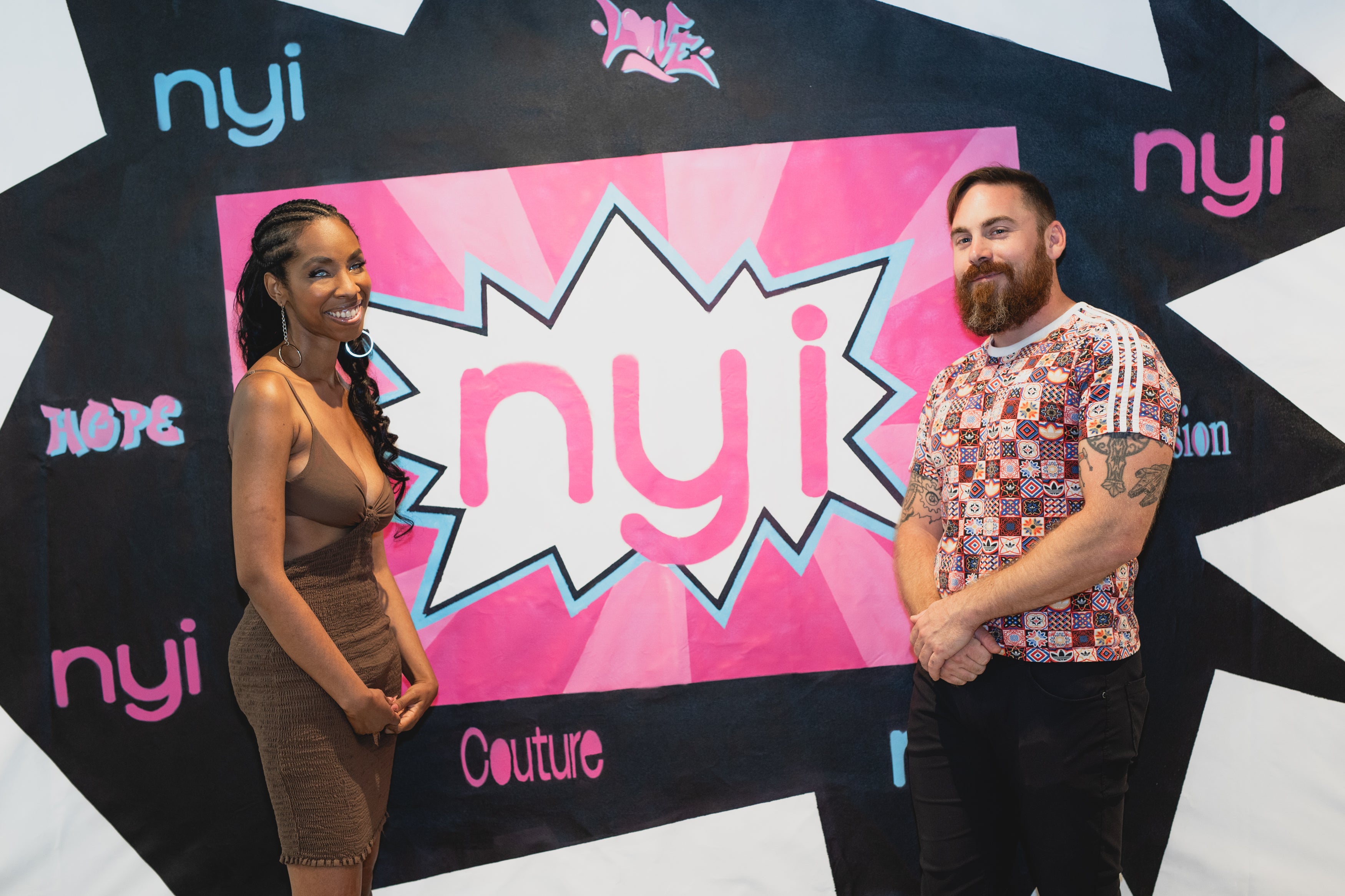 A black woman is standing on the left side front of a pink and blue pop art style backdrop with N Y I in large letters. She is wearing a brown dress, large hoop earrings, and high heels with her hand on her hip. A white man with a red beard and short brown hair wearing a multicolored shirt with black pants stands to the right.