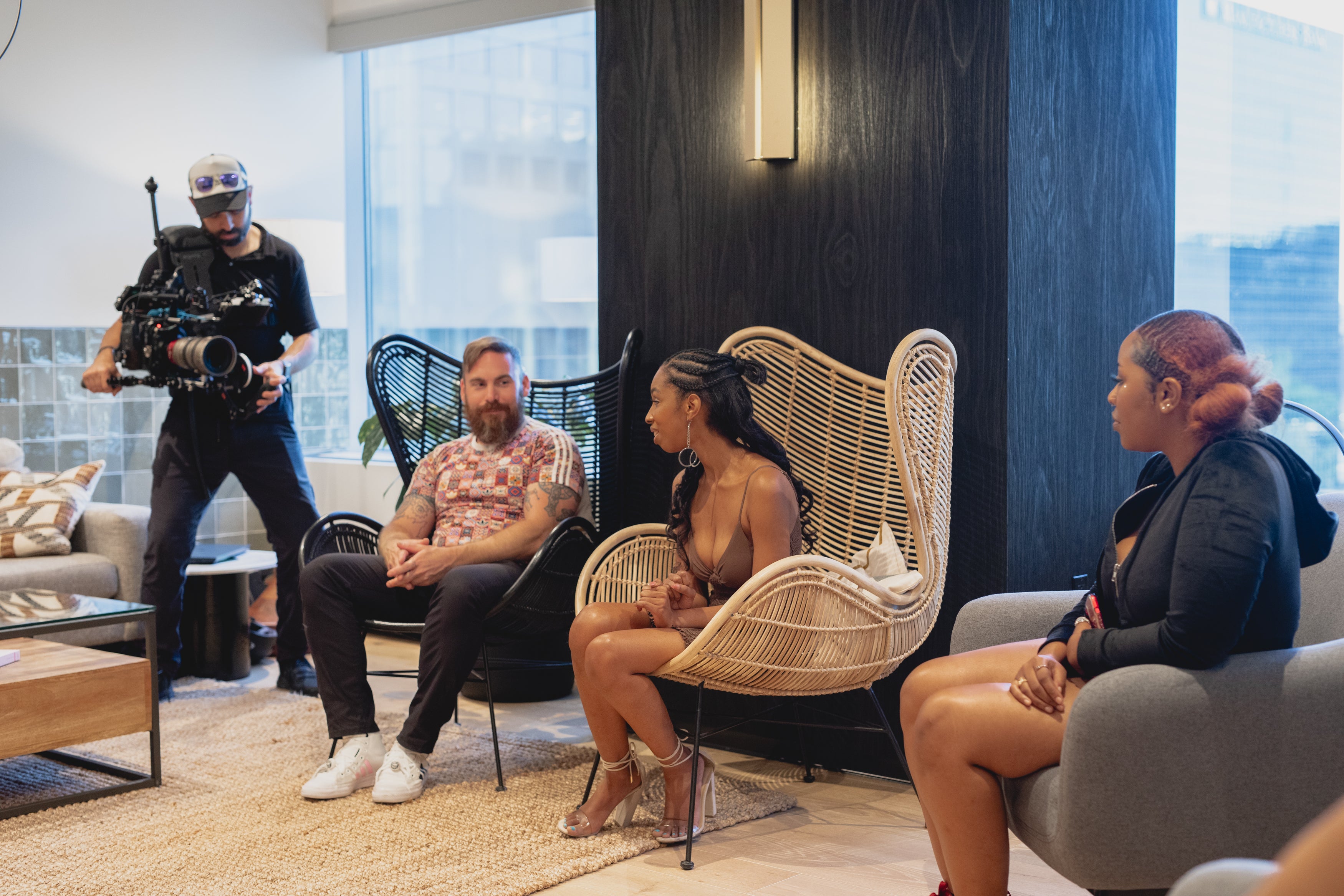 A black woman with long dark hair, wearing a brown dress and high heels sits in a large white wicker chair answering questions from a black woman with short red hair to the right.  A white man with a red beard and short blonde hair sits to the left in a black wicker chair also talking.  A white man stands to the far left holding a camera to record the event.