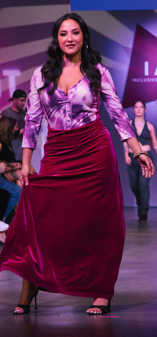 A white woman with long dark hair wearing large gold hoop earings and two piece set that includes a fuchsia, light pink, white, black tie-dye pattern long sleeve bodysuit with a low V-neck line and buttons going down the middle. It’s paired with a fuchsia velvet high slit skirt.  She is wearing brown high heels posing at the end of a runway.
