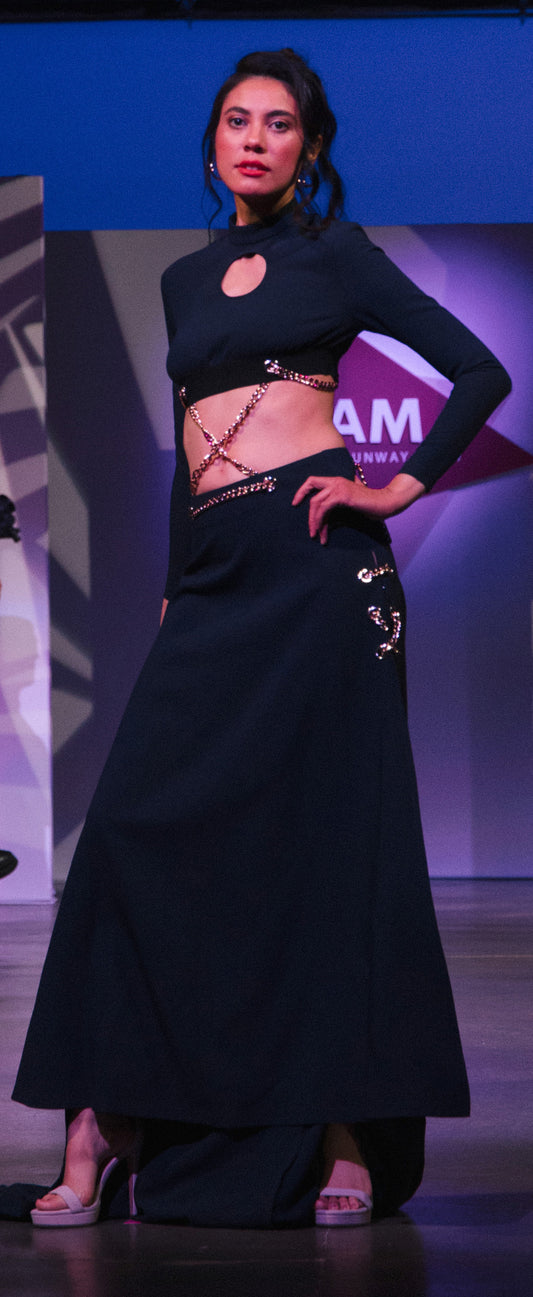 A white woman with long dark hair wears a navy blue two-piece dress that includes a long sleeve crop top with a front cut out made of a suited material. It comes with a matching skirt with a high slit and long train. The top and skirt are connected by a series of gold chains that crisscross to make it one dress.  She is posing with one hand on her hip at the beginning of a pink lit runway.