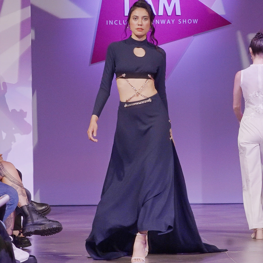 A white woman with long dark hair wears a navy blue two-piece dress that includes a long sleeve crop top with a front cut out made of a suited material. It comes with a matching skirt with a high slit and long train. The top and skirt are connected by a series of gold chains that crisscross to make it one dress.  She is walking confidently down a pink lit runway wearing nude colored high heels with the dress having an 18 inch train flaring behind her.
